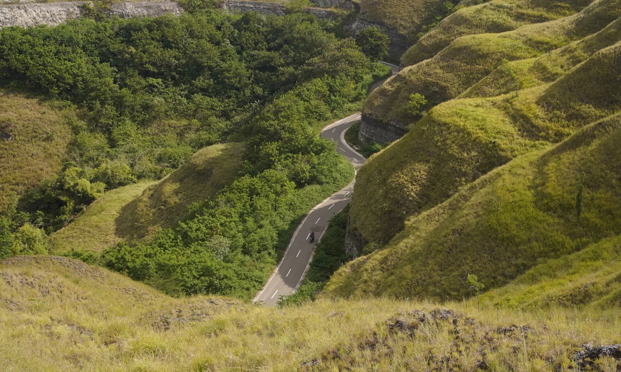 Jalan beraspal berkelok menuruni perbukitan Sumba, dengan lembah di kejauhan.